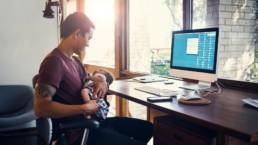 Aikuinen istuu tietokoneen ääressä ja pitää sylissään pientä vauvaa.